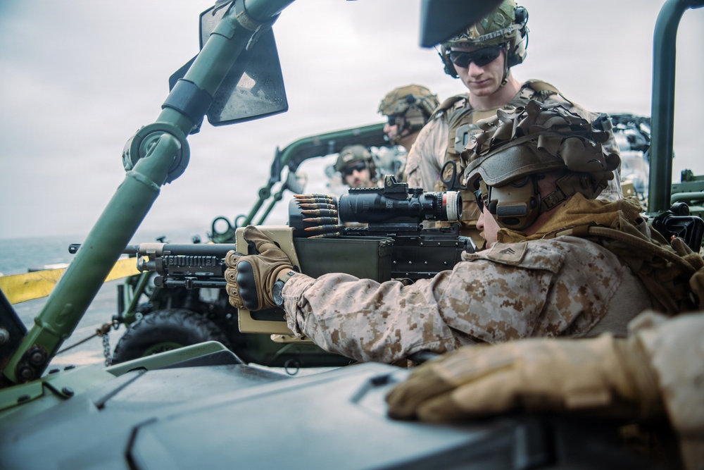 Military personnel, appearing to be U.S. Marines based on their MARPAT digital camouflage uniforms, are positioned on or around a military vehicle, likely a Humvee or similar tactical vehicle. The foreground soldier is manning what appears to be an M240 or similar belt-fed machine gun equipped with an optic sight. The setting appears to be an open-air maritime or coastal environment based on the visible horizon and overcast sky.