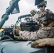 Military personnel, appearing to be U.S. Marines based on their MARPAT digital camouflage uniforms, are positioned on or around a military vehicle, likely a Humvee or similar tactical vehicle. The foreground soldier is manning what appears to be an M240 or similar belt-fed machine gun equipped with an optic sight. The setting appears to be an open-air maritime or coastal environment based on the visible horizon and overcast sky.