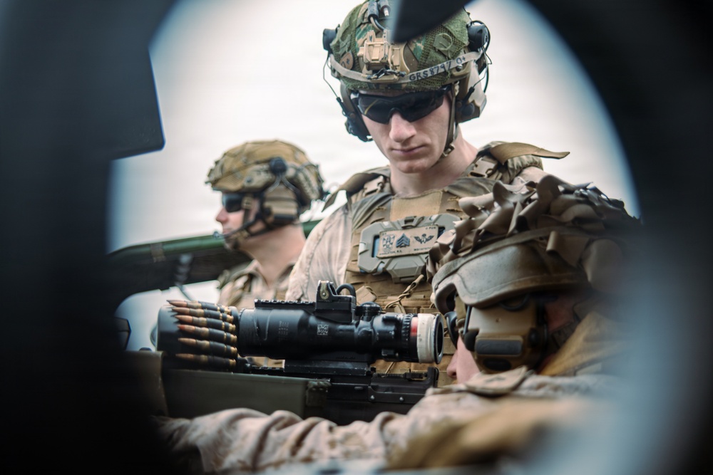 The image shows multiple military personnel in full combat gear, including helmets, tactical vests, and communications equipment. They appear to be positioned around a crew-served weapon system with an optical scope and ammunition belt visible. The photo is taken from a tight angle, likely through or around vehicle or equipment aperture.
