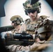 The image shows multiple military personnel in full combat gear, including helmets, tactical vests, and communications equipment. They appear to be positioned around a crew-served weapon system with an optical scope and ammunition belt visible. The photo is taken from a tight angle, likely through or around vehicle or equipment aperture.