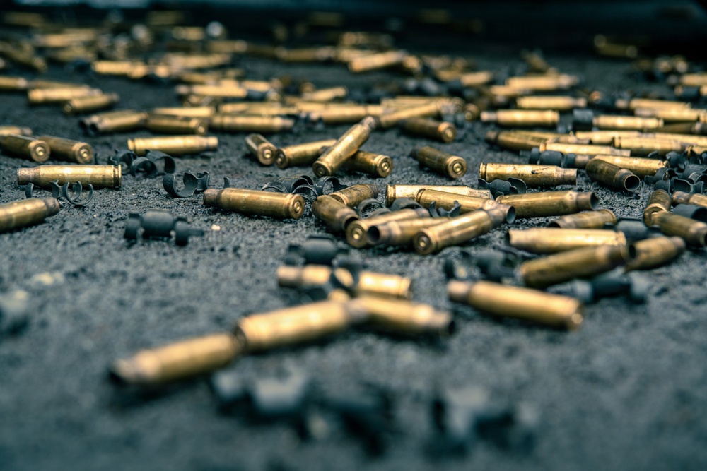 The image shows a large number of spent brass cartridge casings scattered across a dark, gritty surface. Mixed among the casings are deformed metal fragments, likely bullet jackets or wad cutters from fired projectiles. The scene is consistent with a shooting range or post-firing cleanup area.