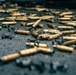 The image shows a large number of spent brass cartridge casings scattered across a dark, gritty surface. Mixed among the casings are deformed metal fragments, likely bullet jackets or wad cutters from fired projectiles. The scene is consistent with a shooting range or post-firing cleanup area.