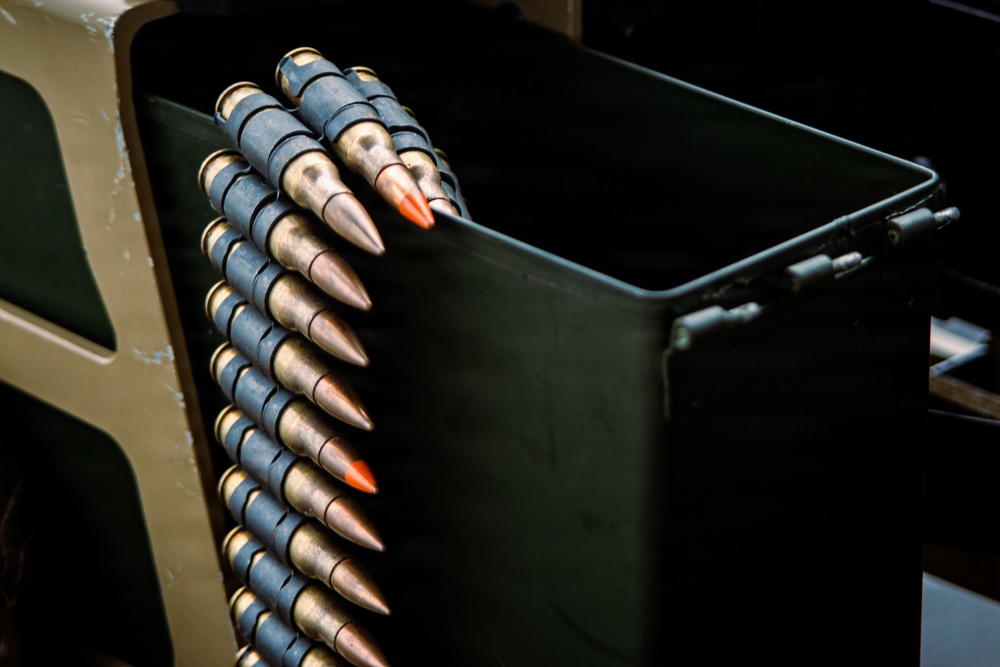 The image shows a belt of linked ammunition rounds draped over the edge of a dark metal ammunition can or box. Several rounds feature distinctive orange-tipped projectiles, indicating tracer or incendiary ammunition. The equipment appears to be associated with a military machine gun or heavy weapon system.