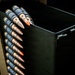 The image shows a belt of linked ammunition rounds draped over the edge of a dark metal ammunition can or box. Several rounds feature distinctive orange-tipped projectiles, indicating tracer or incendiary ammunition. The equipment appears to be associated with a military machine gun or heavy weapon system.