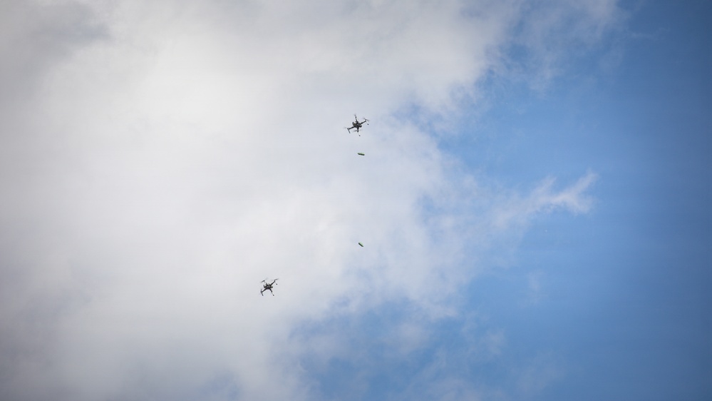 The image shows two multi-rotor drones (quadcopters or hexacopters) photographed from below against a partly cloudy blue sky. Each drone appears to have a small green object suspended or falling beneath it, suggesting a delivery or drop operation. The drones are at different altitudes and horizontal positions within the frame.