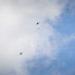 The image shows two multi-rotor drones (quadcopters or hexacopters) photographed from below against a partly cloudy blue sky. Each drone appears to have a small green object suspended or falling beneath it, suggesting a delivery or drop operation. The drones are at different altitudes and horizontal positions within the frame.