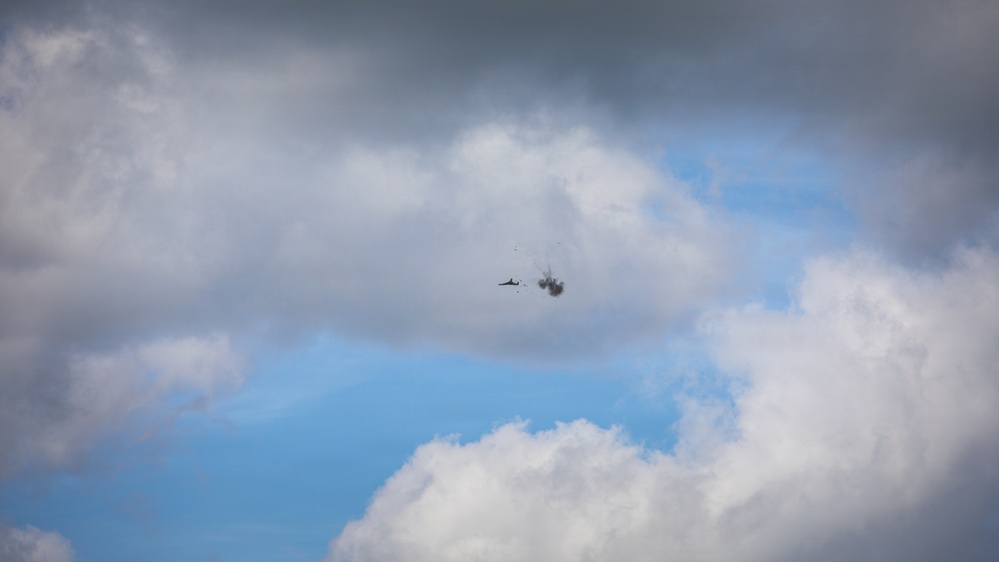 The image shows an aircraft in flight against a partly cloudy blue sky, with a visible explosion or detonation occurring immediately adjacent to or around the aircraft. Debris or fragmentation appears to be dispersing outward from the blast point. The aircraft and explosion are small and distant, centered in the frame.