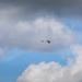 The image shows an aircraft in flight against a partly cloudy blue sky, with a visible explosion or detonation occurring immediately adjacent to or around the aircraft. Debris or fragmentation appears to be dispersing outward from the blast point. The aircraft and explosion are small and distant, centered in the frame.