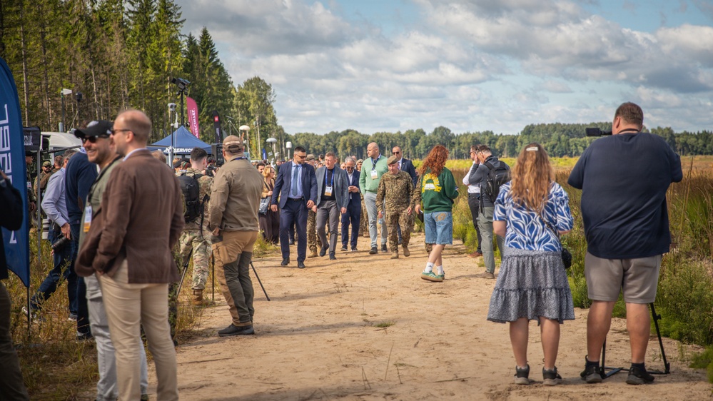 An outdoor event, likely a military or defense exhibition, taking place on a sandy dirt path bordered by forest and open fields. A mixed group of civilians, military personnel in camouflage uniforms, and officials wearing suits and lanyards/badges are walking together, suggesting a guided tour or demonstration. Media personnel with cameras and tripods are present along the sides, documenting the event.
