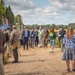 An outdoor event, likely a military or defense exhibition, taking place on a sandy dirt path bordered by forest and open fields. A mixed group of civilians, military personnel in camouflage uniforms, and officials wearing suits and lanyards/badges are walking together, suggesting a guided tour or demonstration. Media personnel with cameras and tripods are present along the sides, documenting the event.