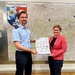 Two individuals are standing together in what appears to be an official police office, jointly holding a document or poster. Behind them is a large map on the wall and an official emblem/crest labeled 'Bundespolizeiinspektion Rosenheim' (Federal Police Inspection Rosenheim). The person on the left is wearing a light blue police uniform, while the person on the right is wearing a pink blazer.