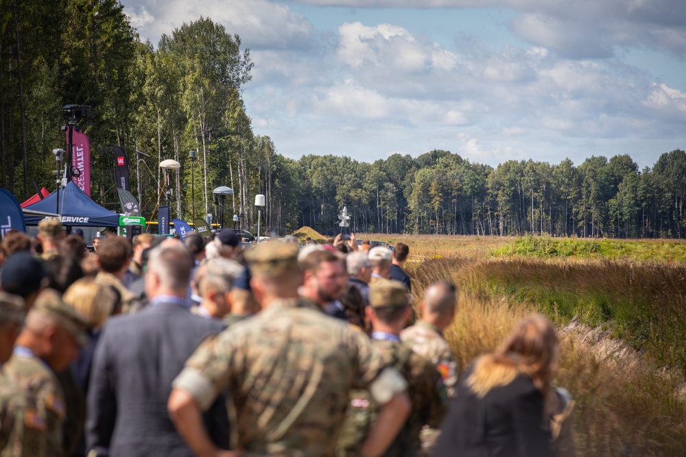 A large outdoor gathering of military personnel and civilians at what appears to be a defense exhibition or military demonstration event. The crowd is positioned along a field edge with vendor tents and equipment visible on the left side. A forested treeline forms the background under a partly cloudy sky.