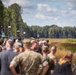 A large outdoor gathering of military personnel and civilians at what appears to be a defense exhibition or military demonstration event. The crowd is positioned along a field edge with vendor tents and equipment visible on the left side. A forested treeline forms the background under a partly cloudy sky.