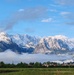 A panoramic landscape photograph showing a snow-capped Alpine mountain range rising dramatically above a low-lying cloud layer. In the foreground, a green meadow and a small village or town with a church spire are visible, surrounded by deciduous and coniferous trees in full leaf. The scene suggests late spring or early summer conditions at lower elevations, with significant snow still present at higher altitudes.