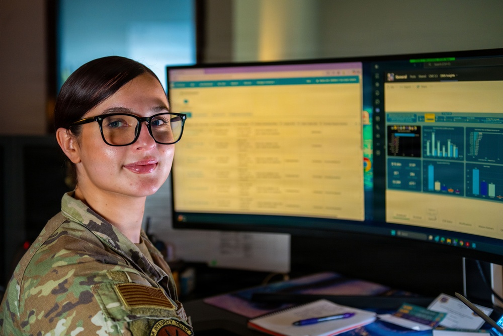 A female military service member in camouflage uniform sits at a dual-monitor workstation, smiling toward the camera. She is wearing black-rimmed glasses and has her hair pulled back. The monitors display what appear to be data dashboards and spreadsheet-style interfaces.