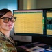 A female military service member in camouflage uniform sits at a dual-monitor workstation, smiling toward the camera. She is wearing black-rimmed glasses and has her hair pulled back. The monitors display what appear to be data dashboards and spreadsheet-style interfaces.