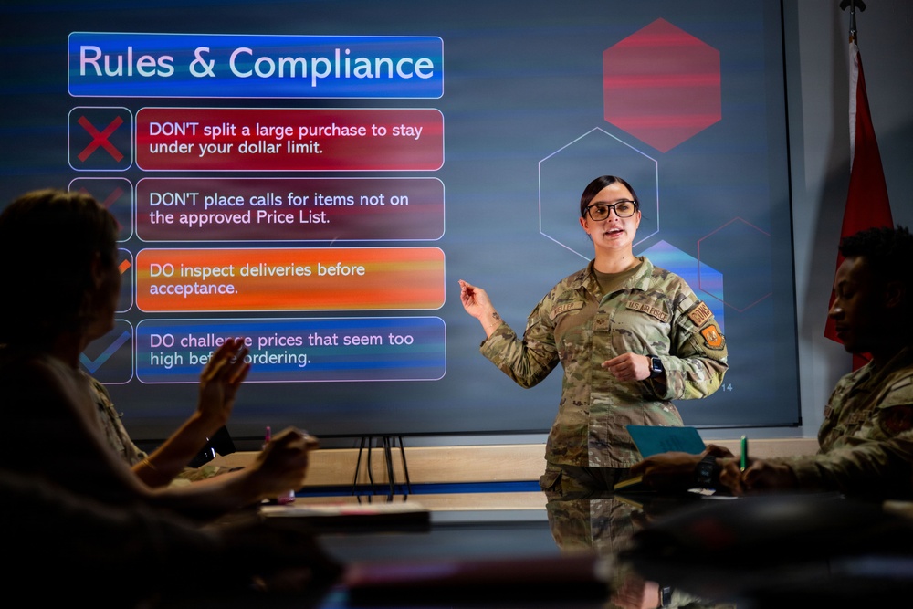 A U.S. Air Force service member is delivering a training or briefing presentation on 'Rules &amp; Compliance' to a small group of military personnel in a conference room setting. The presenter stands beside a large display screen showing procurement or purchasing compliance rules. The audience members are seated at a table, with at least two individuals visible in the foreground.