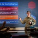 A U.S. Air Force service member is delivering a training or briefing presentation on 'Rules &amp; Compliance' to a small group of military personnel in a conference room setting. The presenter stands beside a large display screen showing procurement or purchasing compliance rules. The audience members are seated at a table, with at least two individuals visible in the foreground.