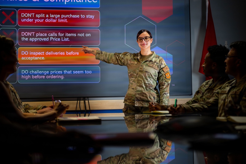 A female military service member in US Air Force OCP (Operational Camouflage Pattern) uniform is conducting a training briefing on procurement rules and compliance. She is pointing at a large display screen showing bullet points about purchasing regulations while addressing seated colleagues at a conference table. The setting appears to be a modern military training or briefing room.