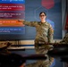 A female military service member in US Air Force OCP (Operational Camouflage Pattern) uniform is conducting a training briefing on procurement rules and compliance. She is pointing at a large display screen showing bullet points about purchasing regulations while addressing seated colleagues at a conference table. The setting appears to be a modern military training or briefing room.