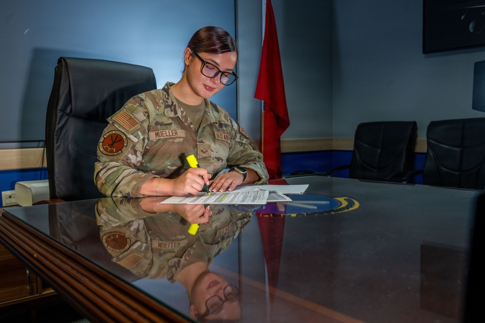 A U.S. Air Force service member named Mueller is seated at a glass-topped conference table, reviewing and highlighting a document. She is wearing the Operational Camouflage Pattern (OCP) uniform with visible name tape and branch insignia. The setting appears to be a formal military office or conference room.