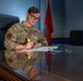 A U.S. Air Force service member named Mueller is seated at a glass-topped conference table, reviewing and highlighting a document. She is wearing the Operational Camouflage Pattern (OCP) uniform with visible name tape and branch insignia. The setting appears to be a formal military office or conference room.