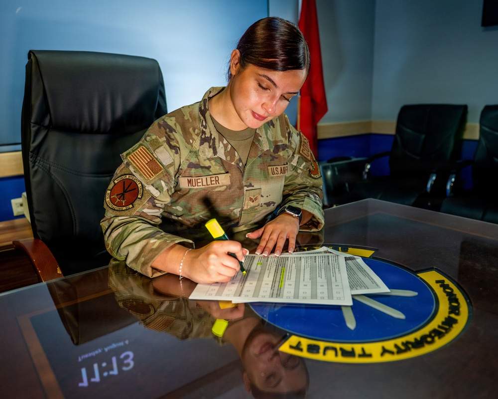 A U.S. Air Force service member named Mueller is seated at a glass-topped conference table, reviewing and highlighting a document. The setting appears to be a formal military office or briefing room, with a unit emblem embedded in the table surface. The emblem visible on the table reads 'HONOR INTEGRITY TRUST' in a circular design.
