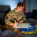 A U.S. Air Force service member named Mueller is seated at a glass-topped conference table, reviewing and highlighting a document. The setting appears to be a formal military office or briefing room, with a unit emblem embedded in the table surface. The emblem visible on the table reads 'HONOR INTEGRITY TRUST' in a circular design.