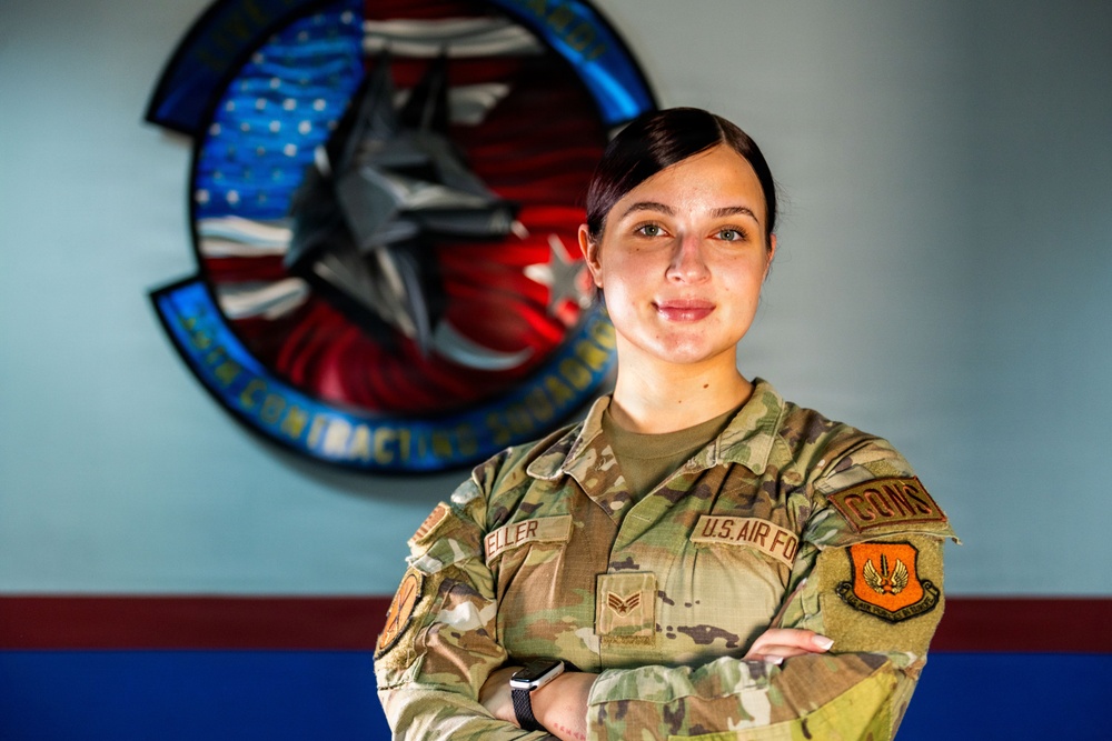A U.S. Air Force service member poses with arms crossed in front of a unit emblem on a wall. The individual is wearing OCP (Operational Camouflage Pattern) uniform with visible name tape reading 'ELLER' and a Senior Airman rank insignia. The background features a squadron patch identified as a Contracting Squadron emblem incorporating an aircraft and American flag elements.