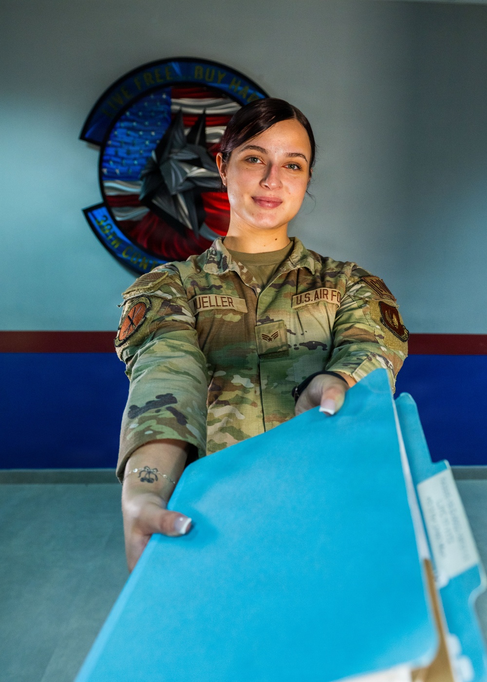 A U.S. Air Force service member wearing OCP (Operational Camouflage Pattern) uniform with the name tape 'MUELLER' and 'U.S. AIR FORCE' tape is extending blue file folders toward the camera. She is smiling and appears to be in an administrative or office setting. A unit patch/emblem is visible on the wall behind her, partially readable as '36th CON...' with red, blue, and American flag design elements.