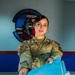 A U.S. Air Force service member wearing OCP (Operational Camouflage Pattern) uniform with the name tape 'MUELLER' and 'U.S. AIR FORCE' tape is extending blue file folders toward the camera. She is smiling and appears to be in an administrative or office setting. A unit patch/emblem is visible on the wall behind her, partially readable as '36th CON...' with red, blue, and American flag design elements.