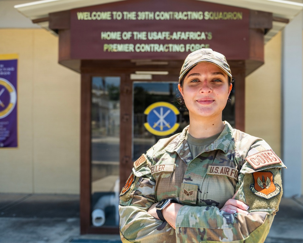 A U.S. Air Force service member stands with arms crossed in front of the 39th Contracting Squadron building. The individual is wearing OCP (Operational Camouflage Pattern) uniform with visible U.S. Air Force tape, a name tape reading 'ELLER', and a 'CONTR' (Contracting) functional badge patch. The building sign identifies it as the home of the USAFE-AFAFRICA Premier Contracting Force.