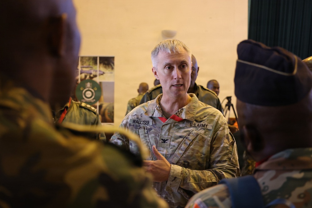 A U.S. Army Colonel named Walliser is engaged in conversation with several foreign military officers in what appears to be an indoor military briefing or meeting environment. He is wearing OCP (Operational Camouflage Pattern) uniform with a Colonel's eagle rank insignia visible on his chest. The setting suggests a multinational military engagement or partnership event.