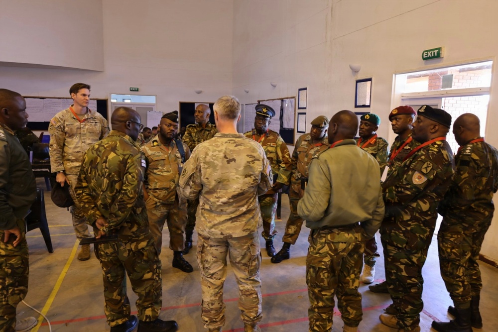 A group of military personnel from multiple nations are gathered in a circle inside an indoor facility, appearing to be engaged in a briefing or discussion. The group includes both Western (likely US) and African military officers wearing various camouflage uniforms and distinctive berets. The setting appears to be a training or conference facility, suggesting a multinational military exercise or cooperation event.
