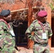 Two military officers in camouflage uniforms are standing outside a red brick building, appearing to be engaged in conversation or observation. The officer on the right wears a maroon beret and has name tapes reading 'MTSETFWA' and 'ESWATINI', indicating affiliation with the Eswatini Defence Force. Both officers are wearing conference lanyards/badges, suggesting attendance at a military event or summit.