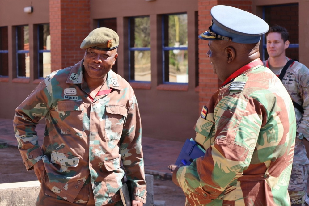 Two military officers in camouflage uniforms are engaged in conversation outdoors in front of a brick building. The officer on the left wears a green beret and has a name tape reading 'MSUTU', while the officer on the right wears a white peaked cap with gold insignia. A third individual in the background appears to be wearing a US Army uniform.