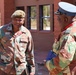 Two military officers in camouflage uniforms are engaged in conversation outdoors in front of a brick building. The officer on the left wears a green beret and has a name tape reading 'MSUTU', while the officer on the right wears a white peaked cap with gold insignia. A third individual in the background appears to be wearing a US Army uniform.
