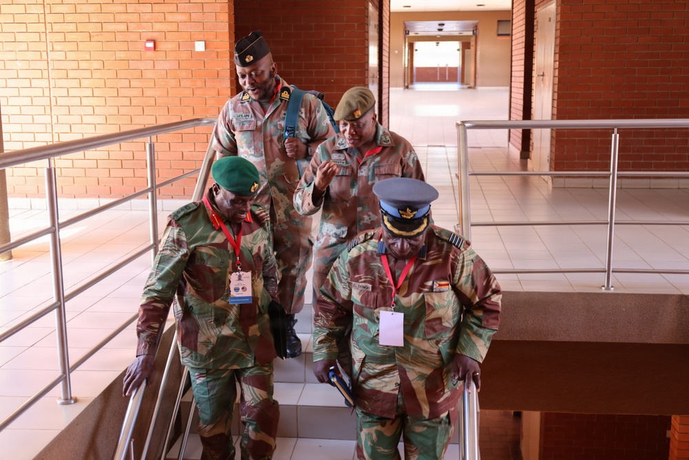 Four military personnel in camouflage uniforms are descending or navigating a staircase inside a brick building. At least two individuals are wearing conference lanyards/badges, suggesting attendance at a formal military meeting or conference. The personnel wear a mix of green berets and a blue peaked cap, indicating possible multi-service or multi-national representation.
