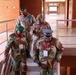 Four military personnel in camouflage uniforms are descending or navigating a staircase inside a brick building. At least two individuals are wearing conference lanyards/badges, suggesting attendance at a formal military meeting or conference. The personnel wear a mix of green berets and a blue peaked cap, indicating possible multi-service or multi-national representation.