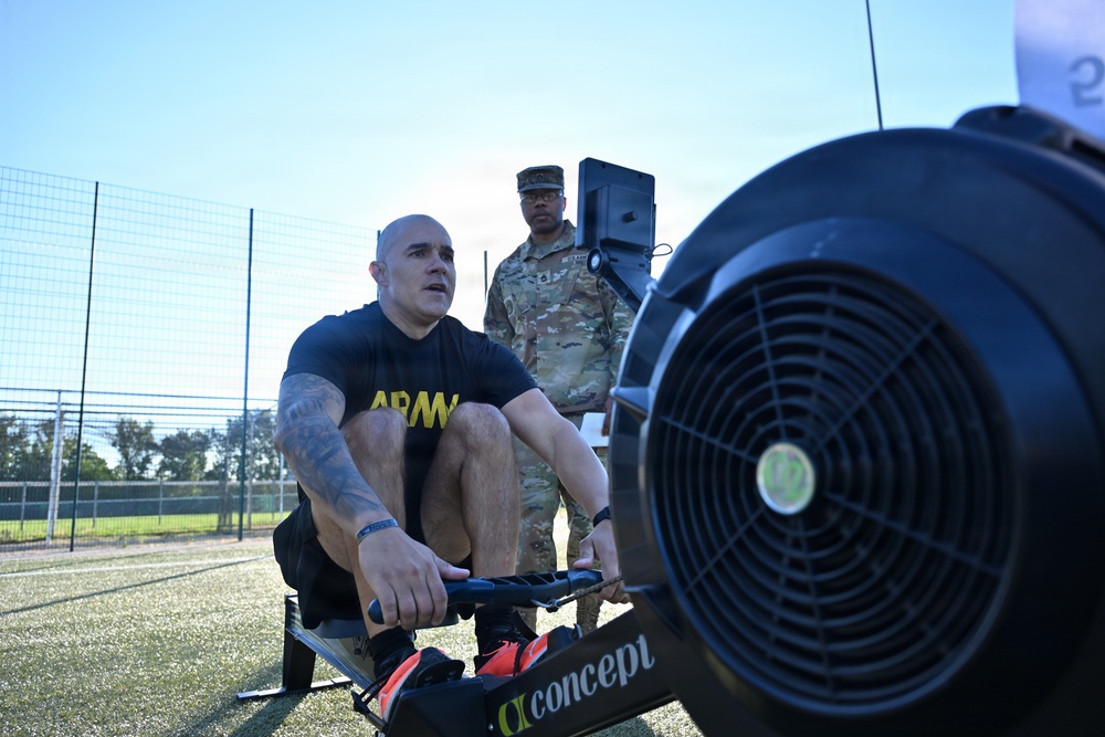A U.S. Army soldier in a black 'ARMY' PT shirt is using a Concept2 rowing machine outdoors on an artificial turf surface. A second soldier in full Army Combat Uniform (ACU/OCP camouflage) stands in the background, appearing to observe or supervise the exercise. The setting appears to be a military physical fitness test or training event.