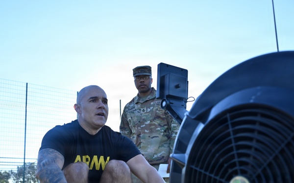 A U.S. Army soldier in a black 'ARMY' PT shirt is using a Concept2 rowing machine outdoors on an artificial turf surface. A second soldier in full Army Combat Uniform (ACU/OCP camouflage) stands in the background, appearing to observe or supervise the exercise. The setting appears to be a military physical fitness test or training event.