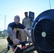 A U.S. Army soldier in a black 'ARMY' PT shirt is using a Concept2 rowing machine outdoors on an artificial turf surface. A second soldier in full Army Combat Uniform (ACU/OCP camouflage) stands in the background, appearing to observe or supervise the exercise. The setting appears to be a military physical fitness test or training event.