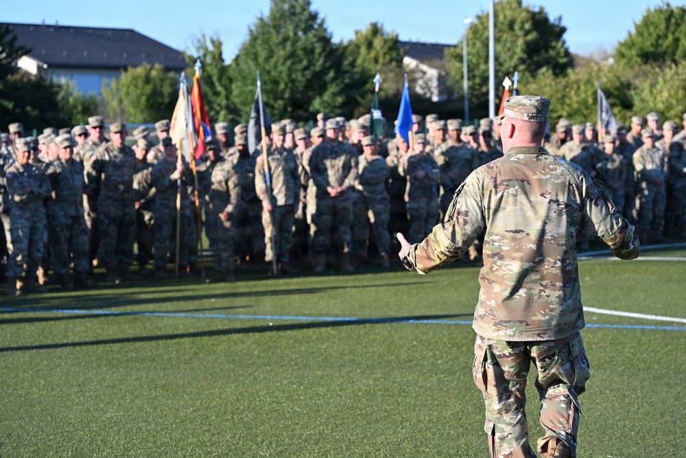 A military officer in OCP (Operational Camouflage Pattern) uniform addresses a large formation of soldiers on an artificial turf field. The speaker stands with his back to the camera, gesturing toward the assembled troops who are standing at ease in organized ranks. Multiple unit guidons and flags are visible among the formation, suggesting a formal military ceremony or briefing.