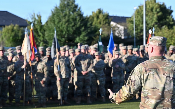 A military officer in OCP (Operational Camouflage Pattern) uniform addresses a large formation of soldiers on an artificial turf field. The speaker stands with his back to the camera, gesturing toward the assembled troops who are standing at ease in organized ranks. Multiple unit guidons and flags are visible among the formation, suggesting a formal military ceremony or briefing.