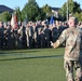 A military officer in OCP (Operational Camouflage Pattern) uniform addresses a large formation of soldiers on an artificial turf field. The speaker stands with his back to the camera, gesturing toward the assembled troops who are standing at ease in organized ranks. Multiple unit guidons and flags are visible among the formation, suggesting a formal military ceremony or briefing.