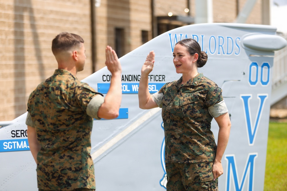 Two military personnel in MARPAT camouflage uniforms are raising their right hands in what appears to be an oath-taking or re-enlistment ceremony. They are standing in front of what appears to be a military aircraft or vehicle marked with 'WARLORDS' and the number '00'. The female service member on the right is wearing a U.S. Navy uniform with visible insignia.
