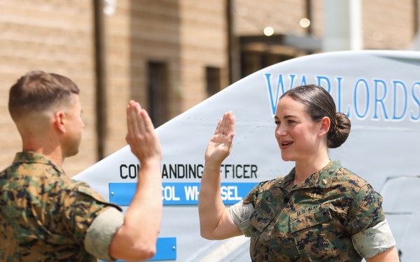 Two military personnel in MARPAT camouflage uniforms are raising their right hands in what appears to be an oath-taking or re-enlistment ceremony. They are standing in front of what appears to be a military aircraft or vehicle marked with 'WARLORDS' and the number '00'. The female service member on the right is wearing a U.S. Navy uniform with visible insignia.