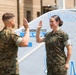 Two military personnel in MARPAT camouflage uniforms are raising their right hands in what appears to be an oath-taking or re-enlistment ceremony. They are standing in front of what appears to be a military aircraft or vehicle marked with 'WARLORDS' and the number '00'. The female service member on the right is wearing a U.S. Navy uniform with visible insignia.