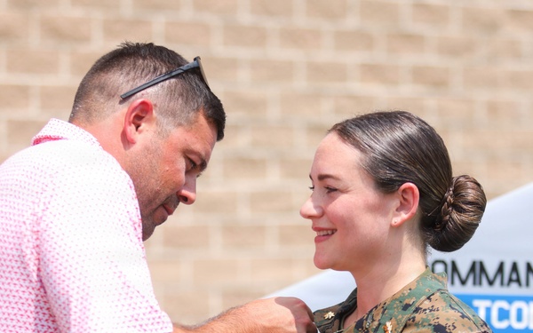 A civilian man in a pink polo shirt is pinning an insignia or device onto the collar/chest area of a female service member in U.S. Marine Corps MARPAT camouflage uniform. The woman is smiling, suggesting this is a celebratory military ceremony such as a promotion or qualification pinning. A sign in the background references a 'Commanding Officer' and partial text including 'LTCOL WHITES' and 'MAJOR DEMAYER', indicating an official military installation setting.