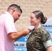 A civilian man in a pink polo shirt is pinning an insignia or device onto the collar/chest area of a female service member in U.S. Marine Corps MARPAT camouflage uniform. The woman is smiling, suggesting this is a celebratory military ceremony such as a promotion or qualification pinning. A sign in the background references a 'Commanding Officer' and partial text including 'LTCOL WHITES' and 'MAJOR DEMAYER', indicating an official military installation setting.